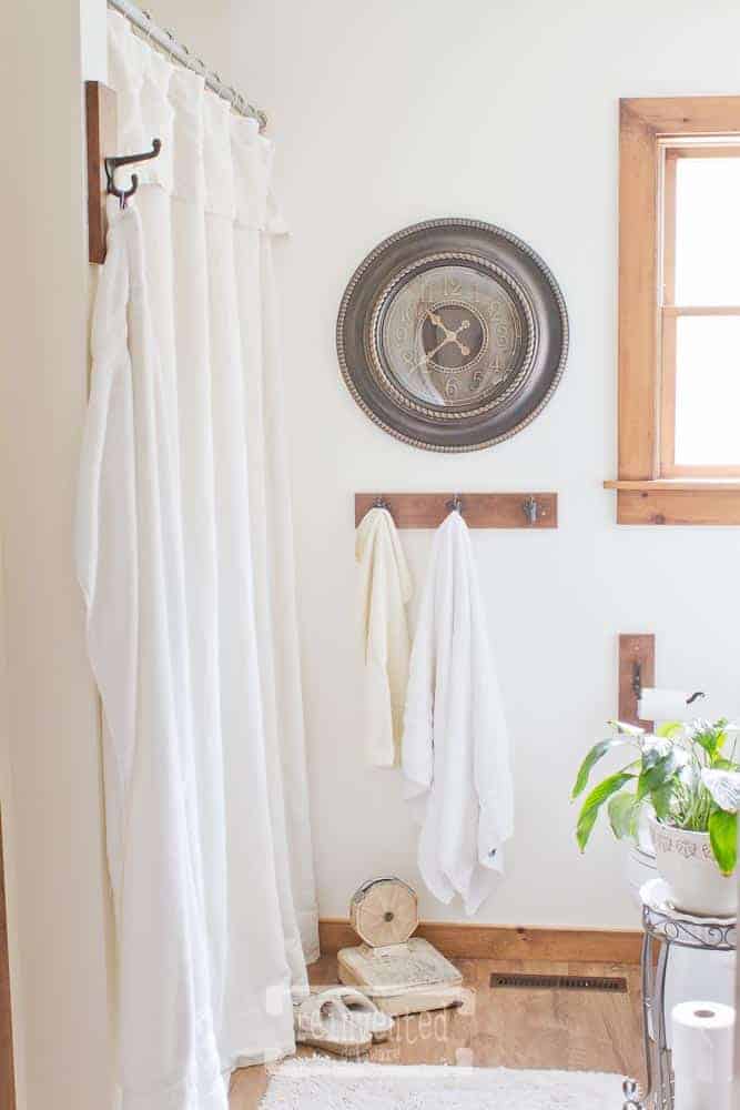 full view of DIY farmhouse shower curtain, clock on the wall, towels hanging on hooks, plant, vintage scales on a vinyl plank floor