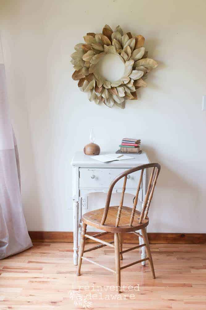 Staged scene showing a sewing cabinet makeover with an antique chair and old books sitting on top with a magnolia wreath hanging on the wall.