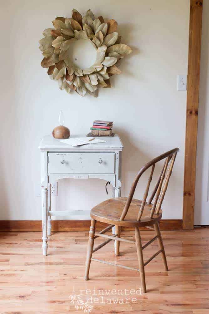 Staged scene with a vintage sewing cabinet that has been milk painted, an old chair, a stack of old books with a writing tablet and a magnolia wreath hanging on the wall.