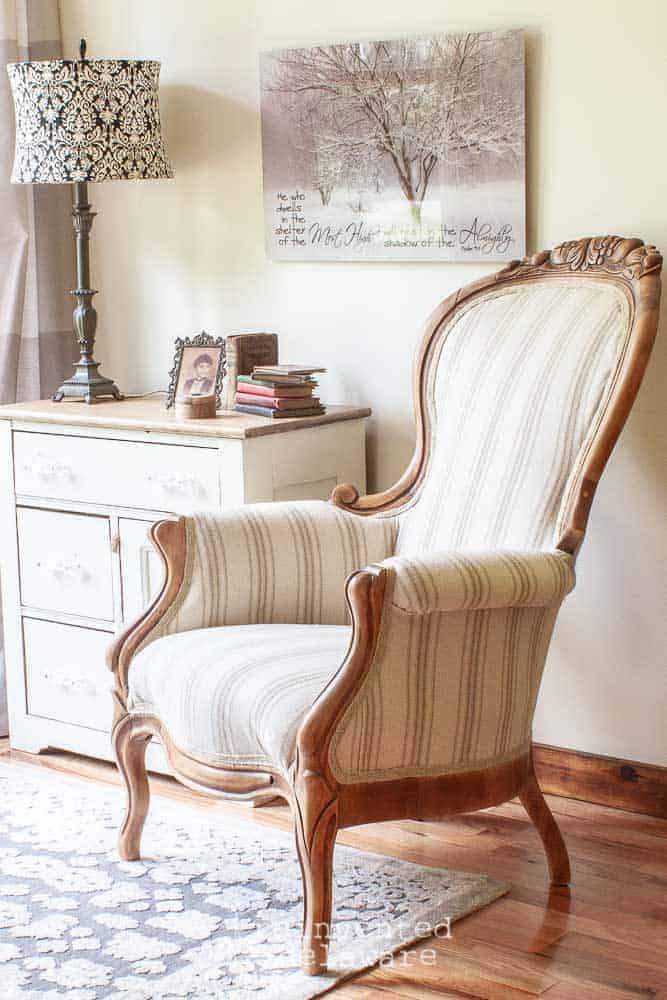 side vidw of Victorian chair that was reupholstered for a blog post tutorial on how to reupholster a chair. Chair is sitting next to a painted washstand with vintage knick-knack sitting on top and a lamp. Wall decor hanging behind the washstand and a gray rug is on the floor