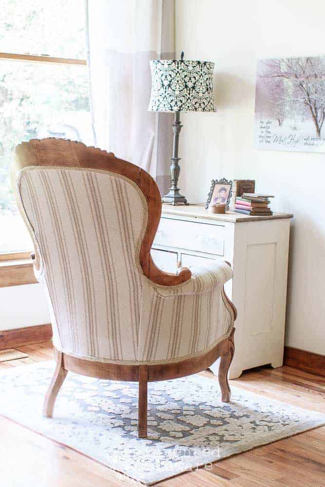 back view of Victorian chair that has been reupholstered in reproduction grainsack fabric in neutral colors. The chair is sitting on a gray rug and in front of a painted antque washstand with knick-knacks and a lamp. Wall decor is hanging on the wall