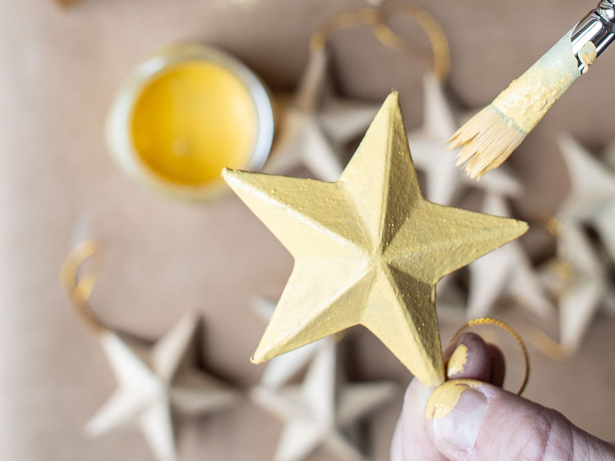 A person holds a golden star ornament, crafted from paper mache, as it's being painted with a brush. In the background, more star ornaments and a jar of golden paint sit on the table, showcasing creative paper mache ideas.