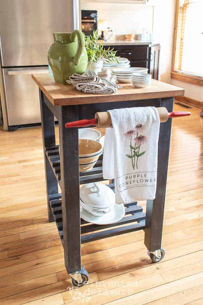 Small kitchen storage cart makeover with rolling pin towel holder and various ironston dishes sitting on top with a Magolia Store Tea Towel hanging.