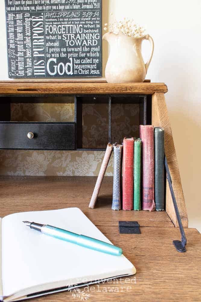 The interior of antique oak fold down desk that has been restored. Stacks of vintage books and a notebook are sititng on top. An ironstone pitcher is sitting on the shelf.