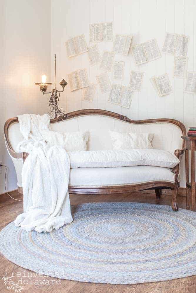 A staged antique sofa with a blue braided rug and book pages stuck on the wall in the background as wall decor.