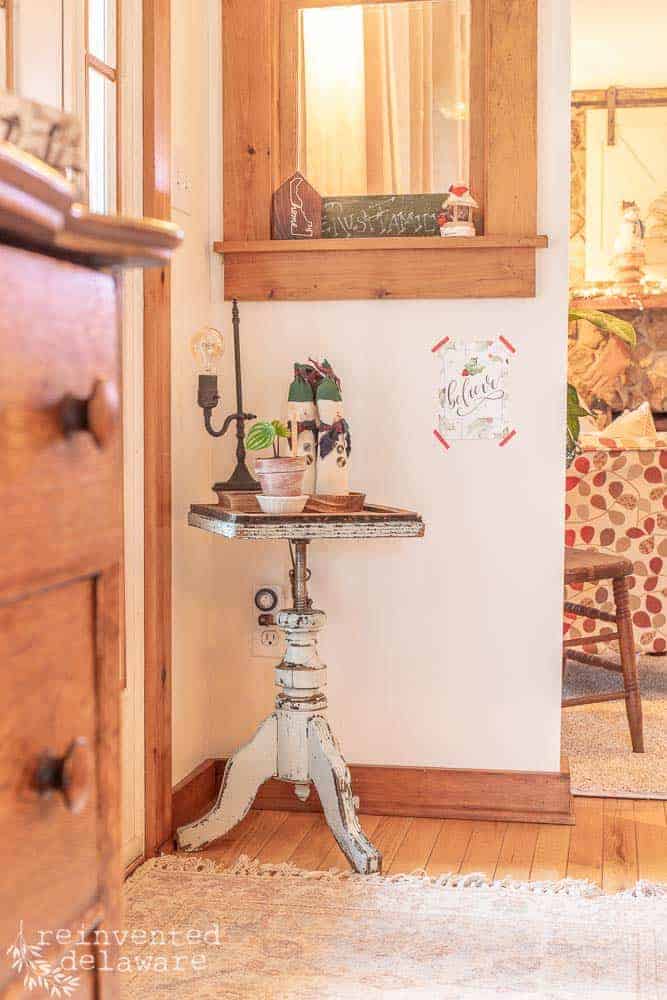 front entryway corner with vintage piano stool as a table with handmade snowmen, plant and little lamp on top