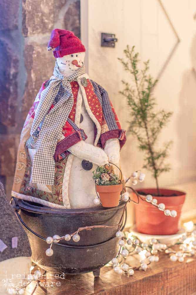 handmade snowman in quilted jacket holding terra cotta pot of pine cones