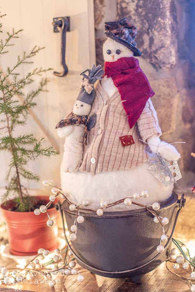 handmade snowman sitting on top of mantle in a vintage cast iron pot. snowman is dressed in upcycle seater hat and jacket