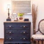 wide shot of gentlemen's dresser that has been painted in Miss Mustard Seed Milk Paint in Typewriter black staged with a lamp, old books, a plant, a chalkboard on the wall, a stack of branches and a chair