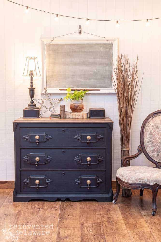 wide shot of gentlemen's dresser that has been painted in Miss Mustard Seed Milk Paint in Typewriter black staged with a lamp, old books, a plant, a chalkboard on the wall, a stack of branches and a chair