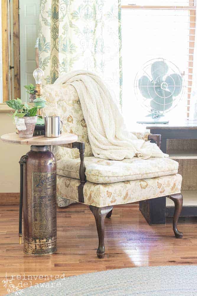 vintage upcycle fire extinguisher side table staged with various items on top and sitting next to a vintage side chair