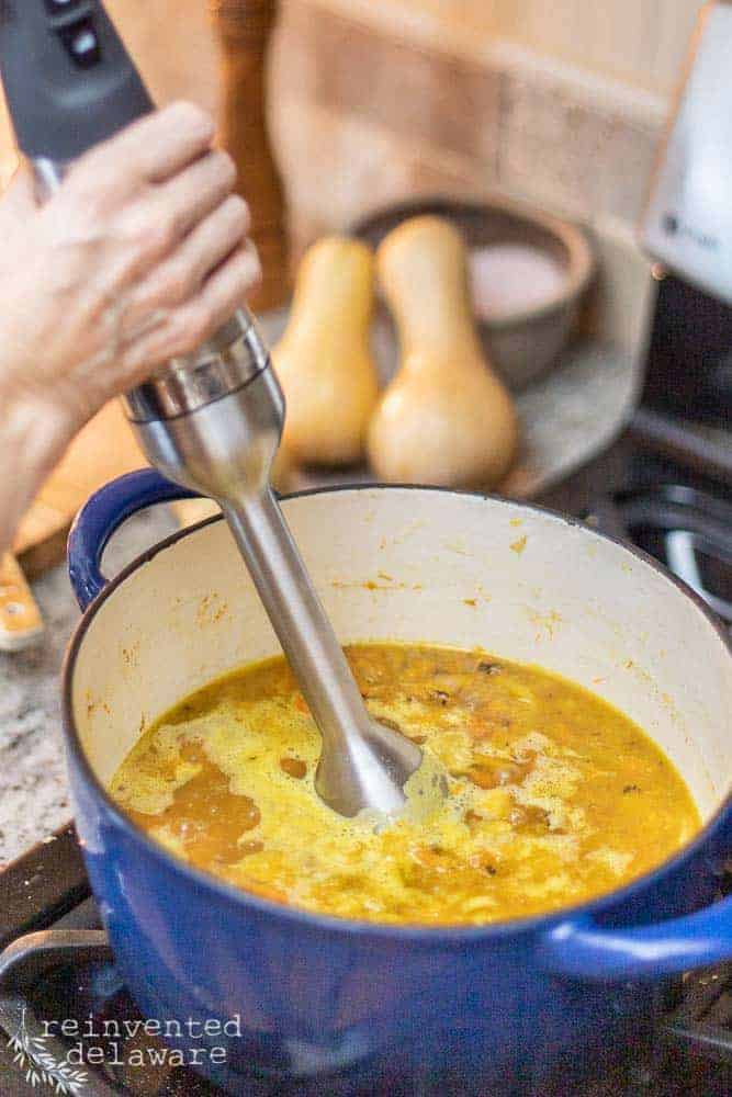 lady using emersion blender in dutch oven filld with butternut squash soup ingredients