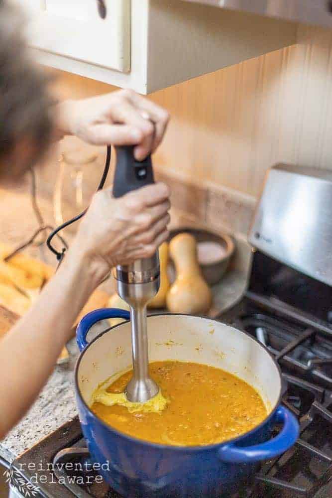 lady using emersion blender in dutch oven filled with butternut squash soup ingredients