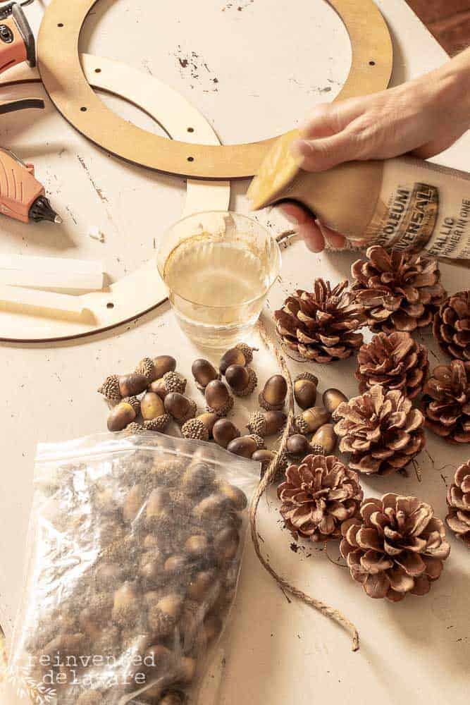 supplies for diy pine cone wreath with acorns laying on work table and lady spraying gold spray paint into a cup of water