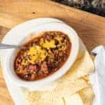 a bowl of chili with chips on a plate, napkin on the side all sitting on a cutting board