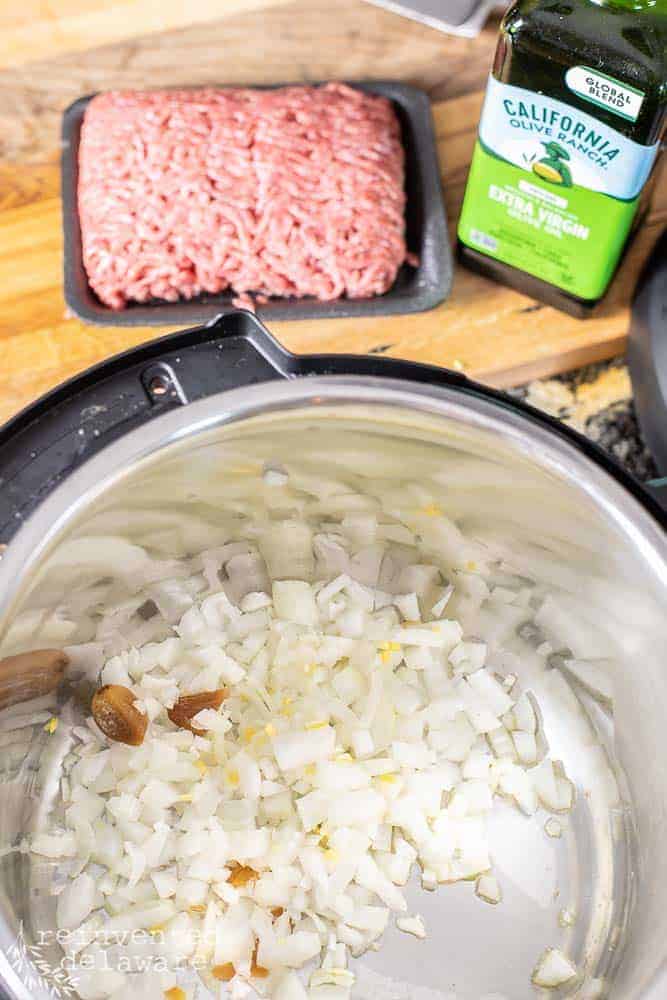 chopped onions and garlic in an Instant Pot sauteing with ground beef and olive oil in background