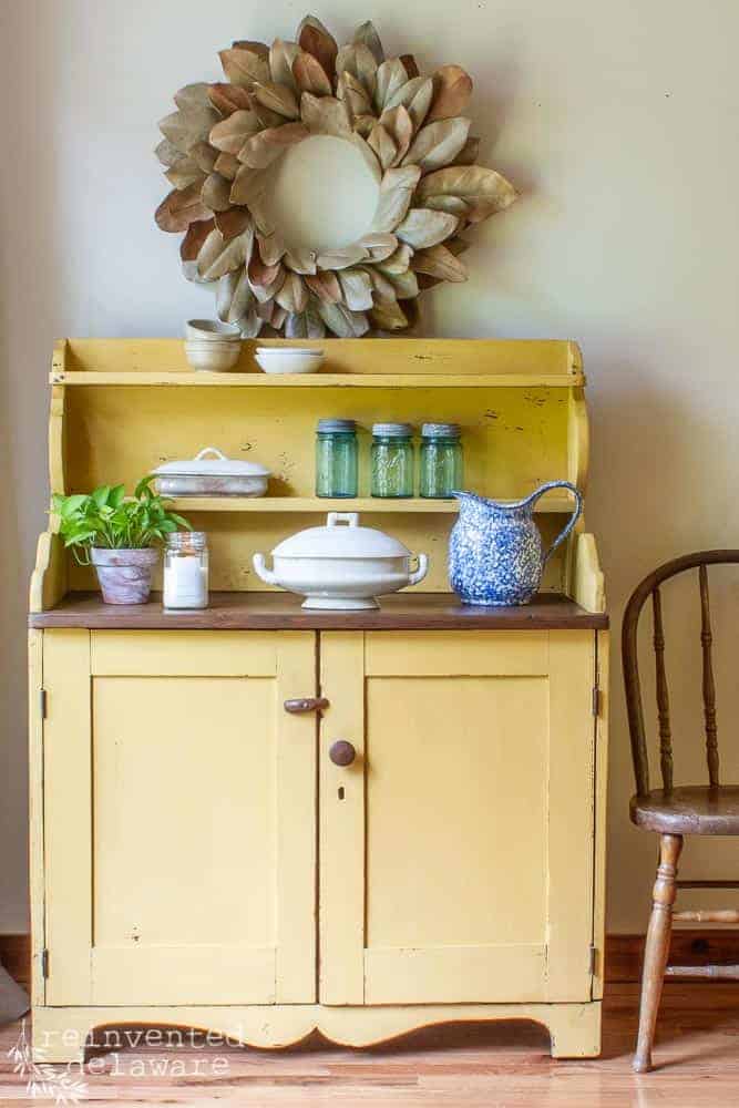 picture of an antique pine cupboard in a tutorial showing how to apply wood paint using milk paint in a yellow color. various items sitting on top of cupboard including a plant, a candle, an ironstone serving dish, a blue pitcher, antique mason jars and several small ironstone and pottery bowls. a magnolia wreath hangs above the painted cabinet
