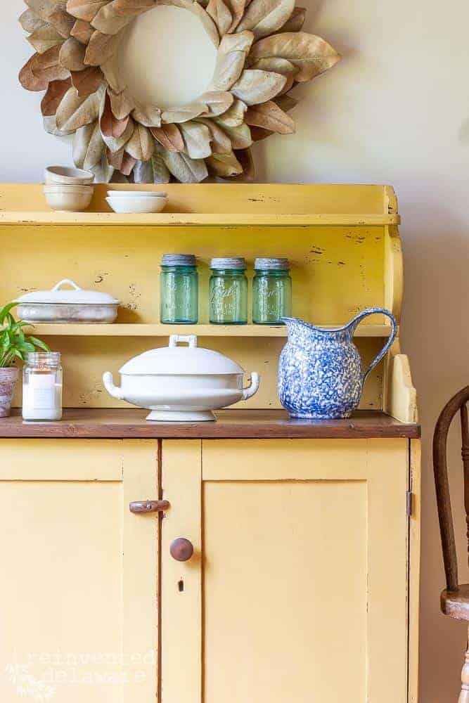 picture of the side of a piece of painted furniture using wood paint in yellow with an ironstone serving bowl, blue pitcher and blue vintage mason jars