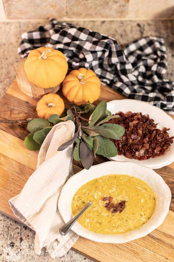 a bowl of chicken corn and butternut squash chowder with bacon bits on the top. a bowl of cooked bacon bits in the background, a sprig of fresh sage, small pumpkins and a buffalo checked towel