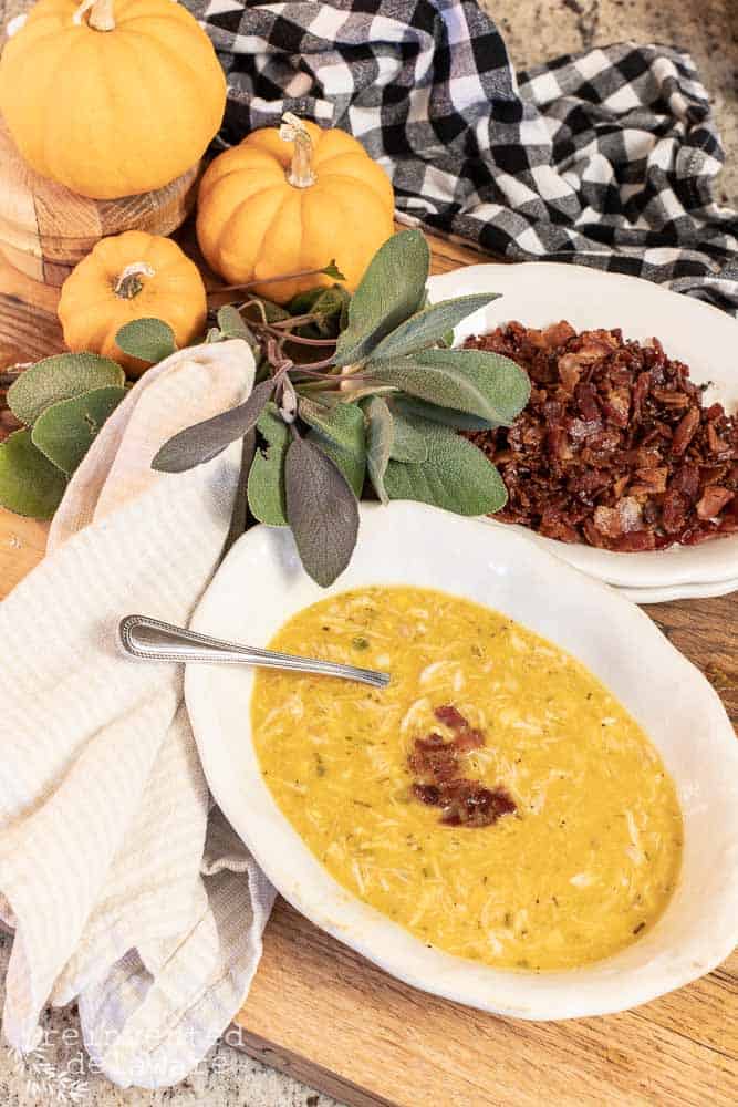 a bowl of chicken corn and butternut squash chowder with bacon on top, a bowl of bacon bits on the side, fresh sage sprigs, small pumpkins and black and white cheched towel in the background.