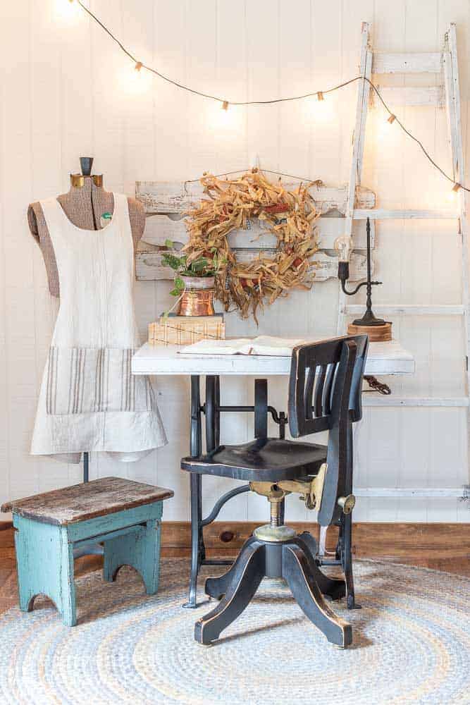 staged corn husk wreath hanging on the wall with a desk, chair, lamp and plant on top of the desk and an antique dress form on the side