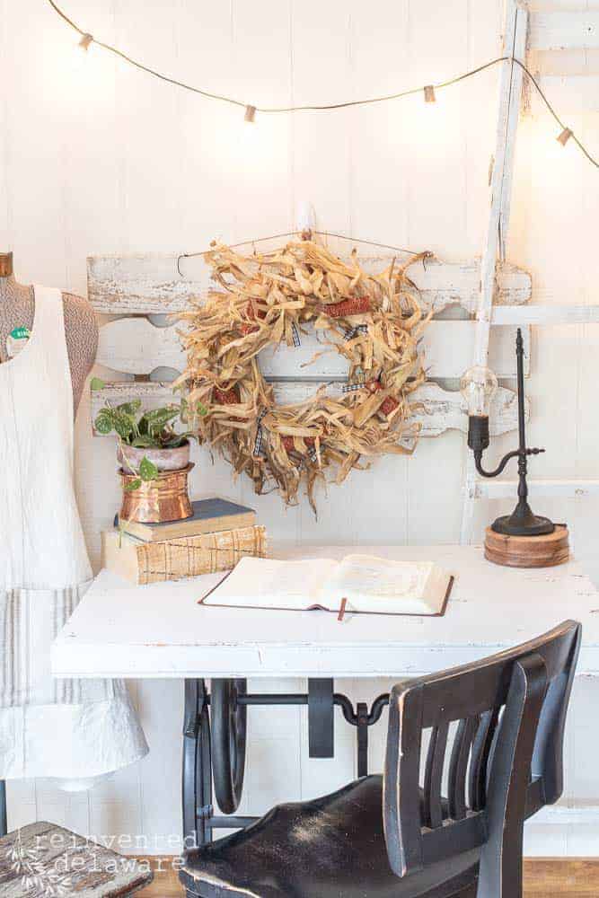 corn husk wreath hanging on a wall with a desk staged with a Bible, a lamp and a plant on top