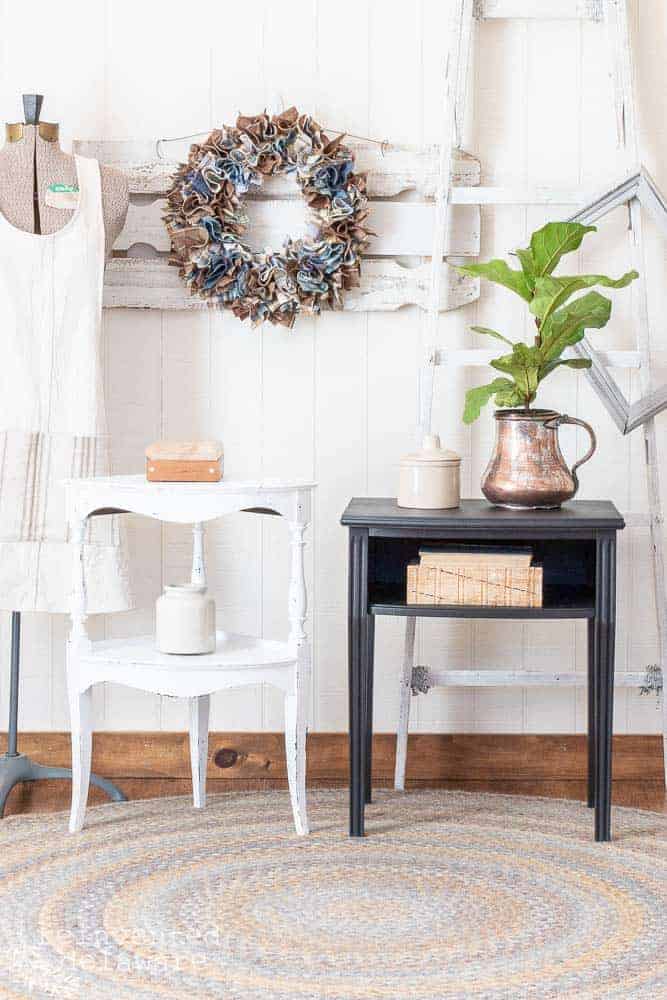 two small tables both with a furniture makeover, one in Dixie Belle Cotton, one in Dixie Belle Midnight Sky for Dixie Belle paint ideas and both are staged with home decor including a wreath on the wall, copper pitcher with a plant, vintage books and a vintage dress form