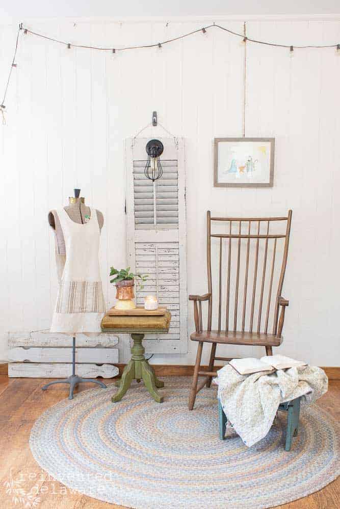 full staged view of old shutter repurposed into a light, wooden chair, dress form with an apron, organ stool, foot stool with a blanket and a Bible on it, wall art and hanging lights in the background