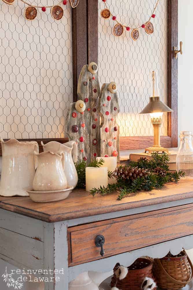 staged Christmas tree home decor piece sitting on top of vintage side board with ironstone, greenery, pine cones and lamp