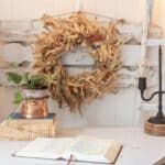 corn husk wreath hanging on the wall behind a desk that has a lamp, a plant and a Bible sitting on top