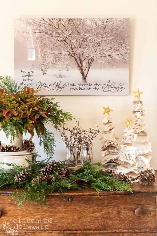 A staged scence showing DIY Farmhouse Christmas trees on a dresser with fresh greenery, faux berries and wall art hanging on the wall.