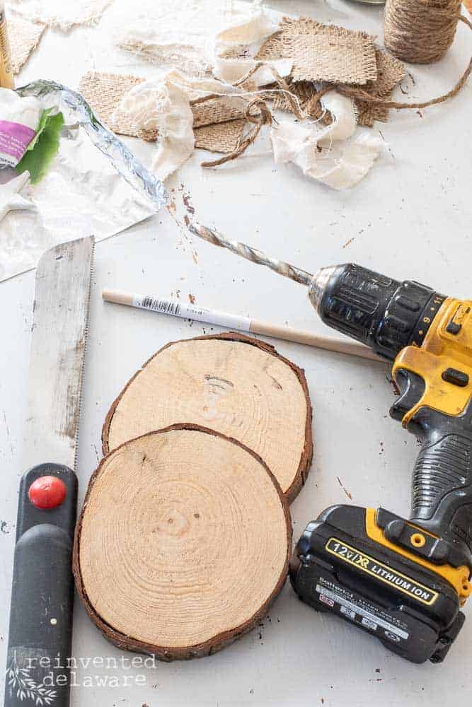 Supplies for DIY holiday Christmas craft including wood slices, power drill, hand saw and fabric scraps.
