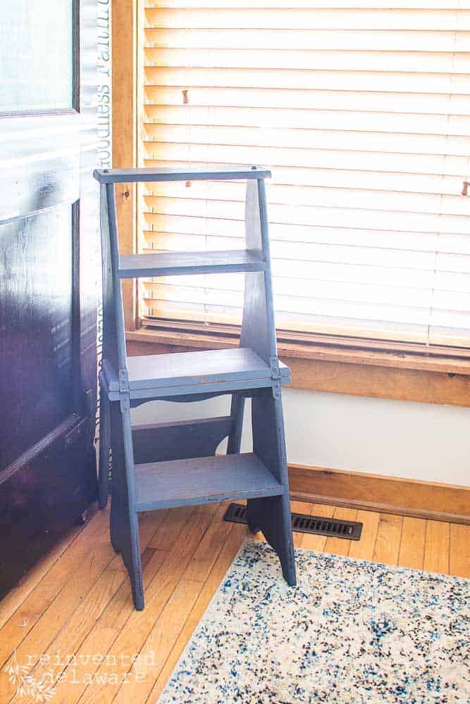 Painted blue foot stool and ladder that converts to a chair sitting in the corner near a window with a blue rug and a brown door.