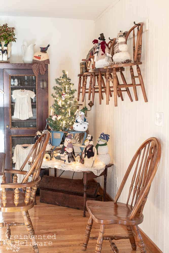 Scene of a dining room with vintage children's chairs hanging on the wall, handmade snowmen on each chair, vintage stroller with various handmade snowmen stacked on it, antique cabinet filled with vintage Pfaltzgraff dishes and childrens dresses hanging on the front of the cabinet.