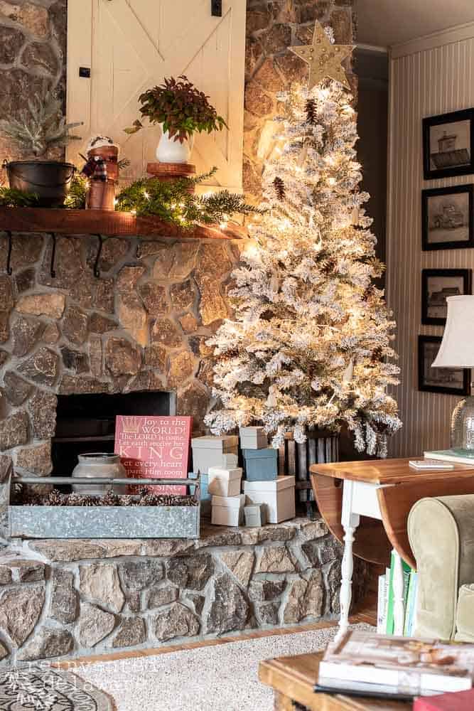 View of a flocked Christmas tree filled with handmade vintage ornaments, painted boxes below, tool caddy filled with pinecones and vintage style Christmas decor on the fireplace mantle.
