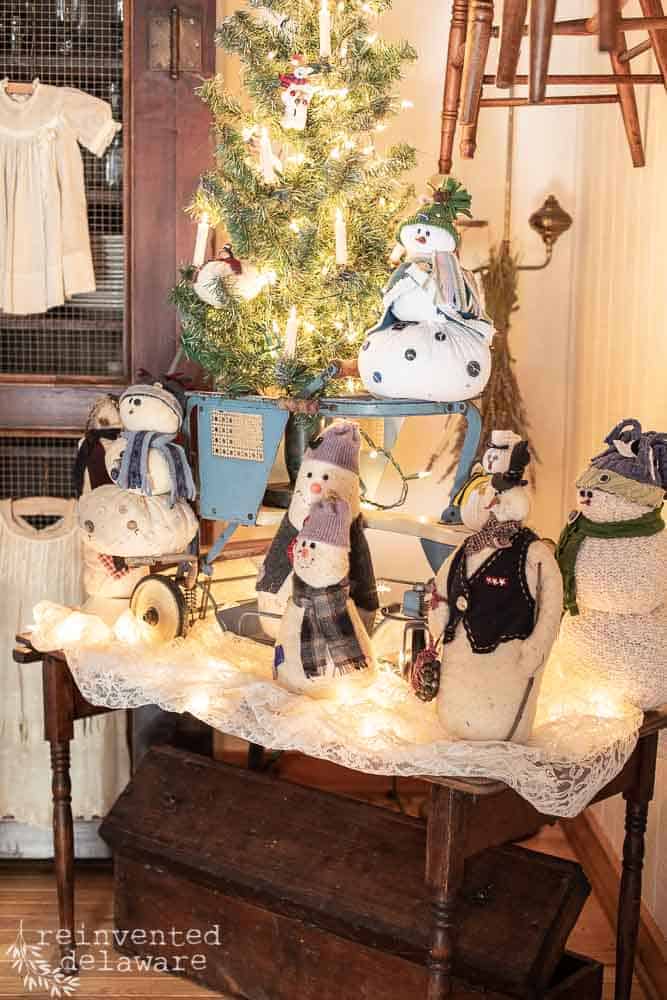 Several handmade snowmen sitting on top of a vintage childs stroller that is on top of an antique sewing table with a vintage wood toolbox underneath.