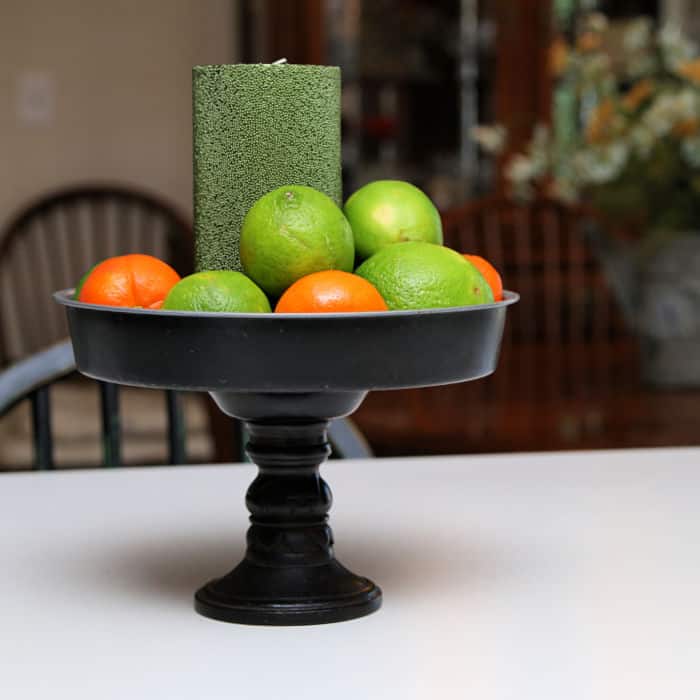 Repurposed thrift store makeover showing a candlestick and a pie pan made into a food riser staged with fruit and a candle.