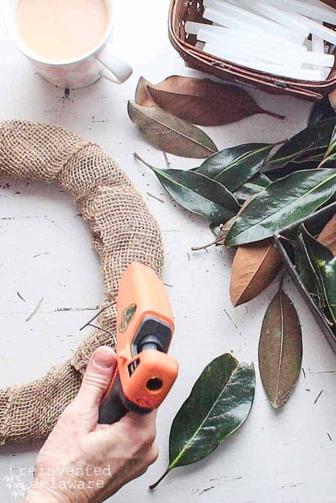Lady using hot glue gun to attach fresh magnolia leaves to a burlap wrapped wire wreath frame with extra leaves, cup of coffee and glue sticks off to the side.
