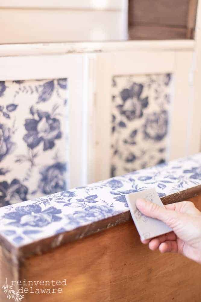 Lady using a scrap of sandpaper to smooth the edges of the decoupage paper for furniture.