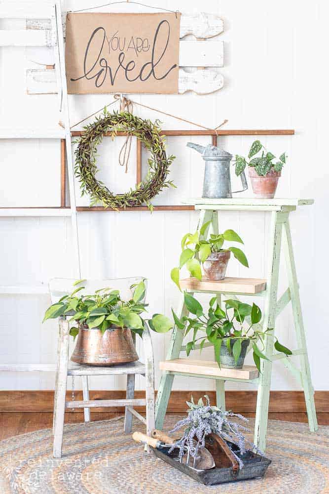 Staged scene showing off an old ladder that was repurposed into a plant stand. Potted plants in terra cotta pots and coppy pots with wall decor help to decorate the scene.