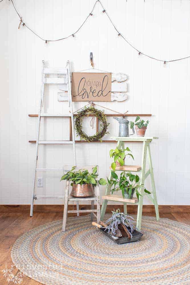 Full front view of a staged scene showing the repurposed step ladder with plants in various containers including a copper pot and terra cotta pots with wall decor in the background.