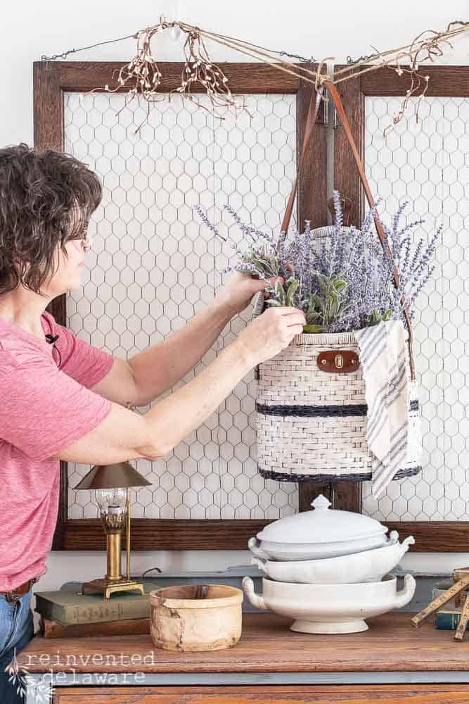 Lady adding faux lavender to a picnic basket upcycle that is hanging as wall decor.