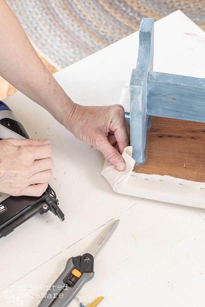 Lady stapling the corners of a footstool makeover with diy grain sack fabric.