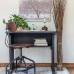 A small upcycled black desk with a brown swivel chair sits on a light rug. The desk holds a plant, books, a candle, and a clock. Wall art of a tree hangs behind it, with tall dried branches arranged to the right.