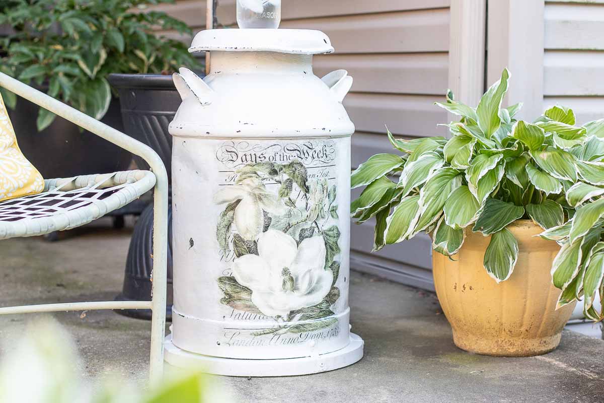 A vintage white milk can with floral designs sits on a patio next to a green-striped potted plant and a metal chair, offering charming inspiration for milk can decorating ideas.
