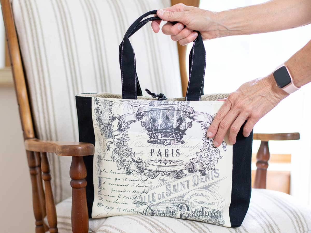A person holds a chic thrift store tote bag with a Paris-themed vintage print and black straps, placed on a wooden armchair with a beige striped cushion. The person's smartwatch glints on their wrist, adding a modern contrast to the upcycled charm of the tote.