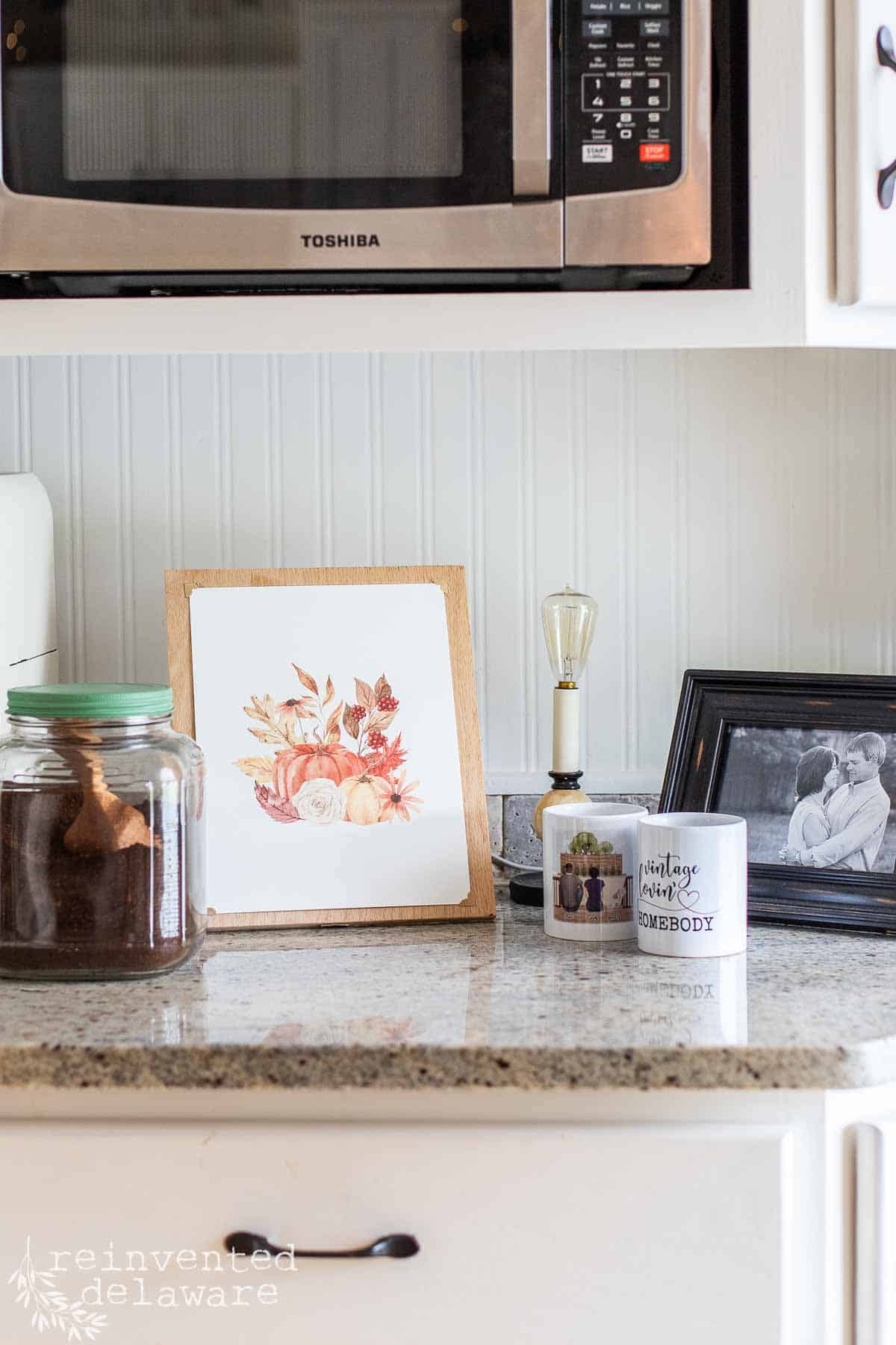 A printable fall decor piece with pumpkins and leaves for fall dislpayed on an easel on a kitchen counter.