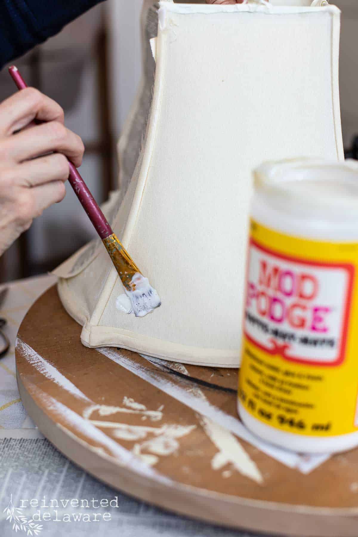 Lady applying Mod Podge to a fabric lampshade for a thrifted lamp makeover project.