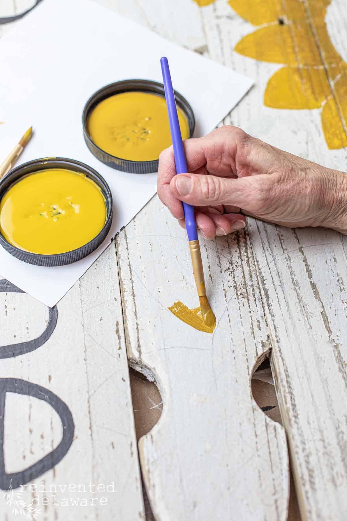 lady using yellow paint to paint a sunflower design on a handmade diy welcome sign for a front door