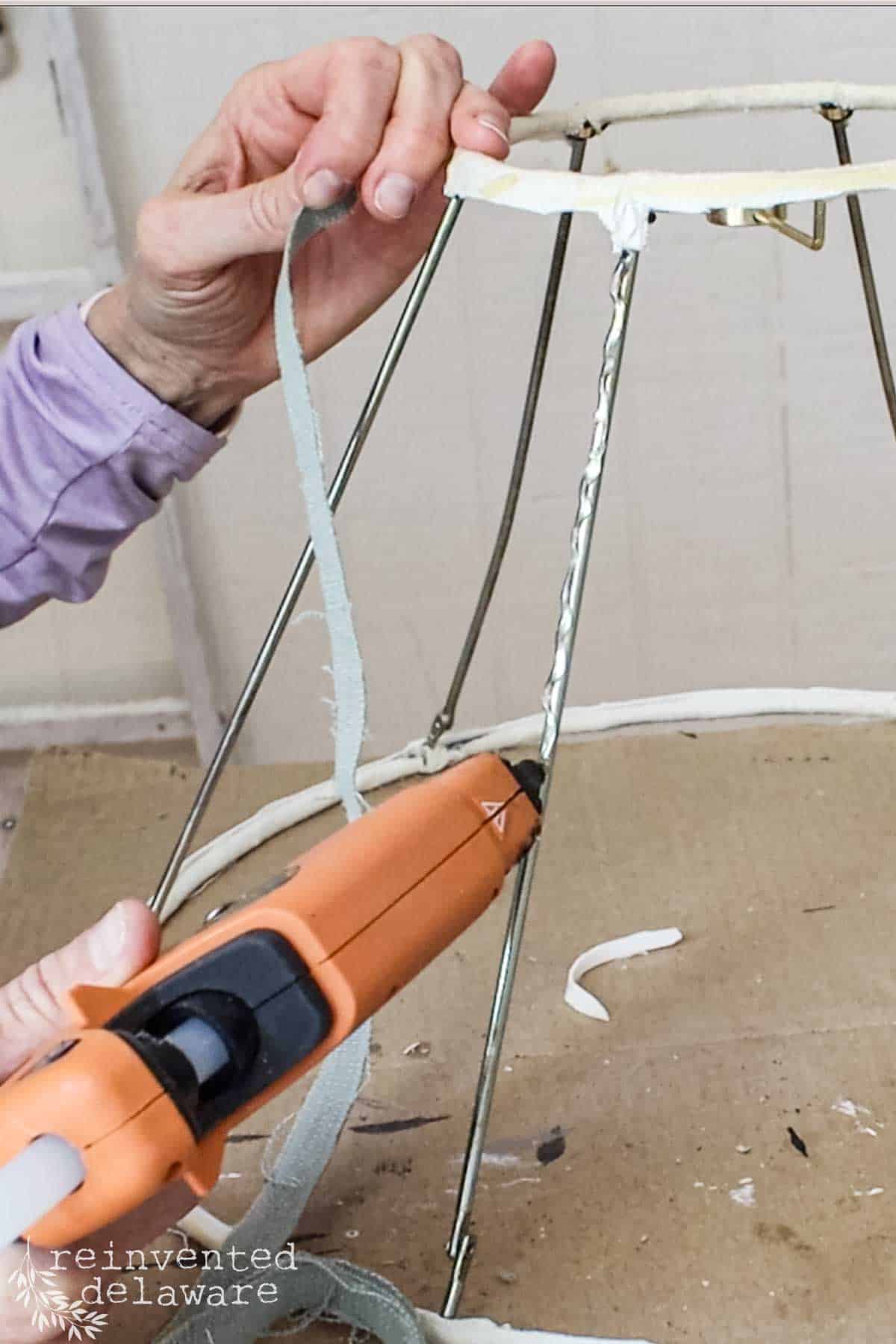 a lady using a glue gun to attach strips of fabric on a wire lampshade for a lampshade makeover hanging light project